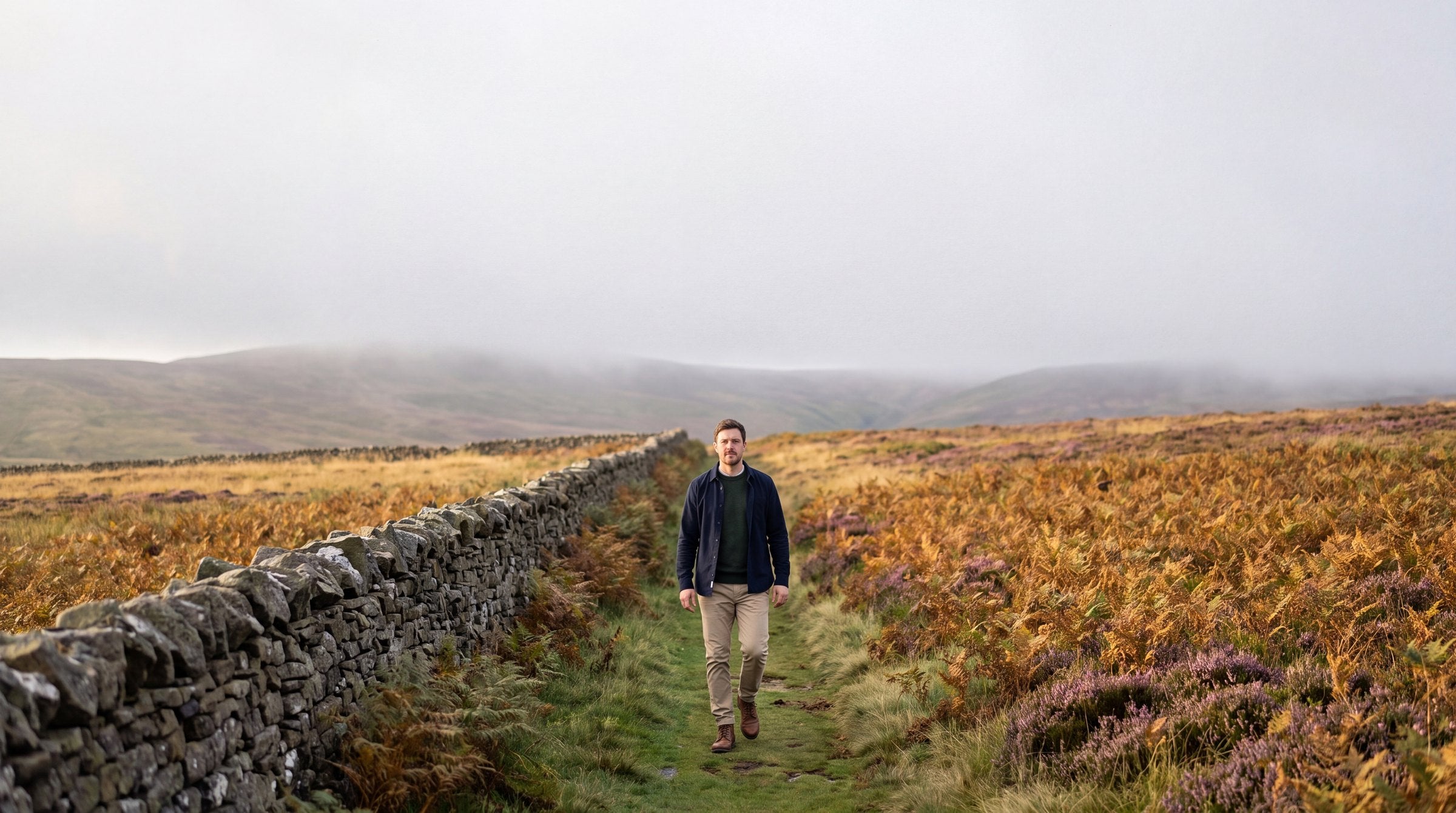 Man walking along a footpath beside a dry stone wall across misty Yorkshire moorland